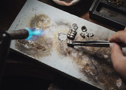 Close-up of a jeweler using a blowtorch to craft silver jewelry pieces on a workbench.
